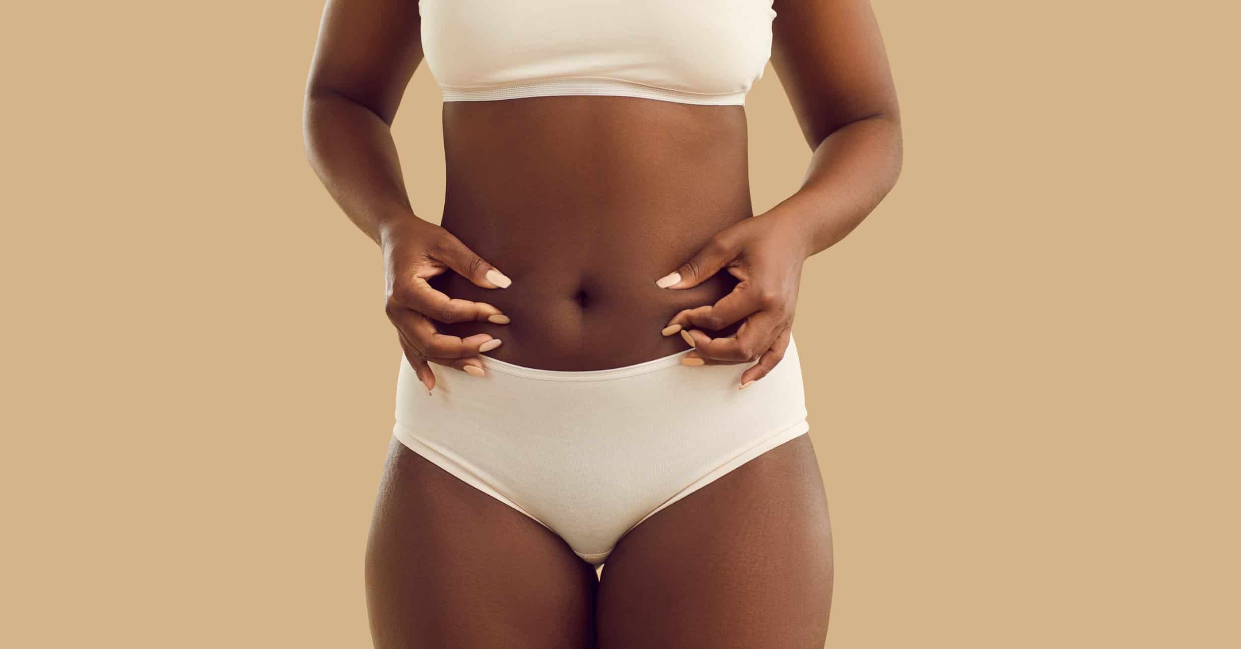 Black lady with some body fat pinches her belly isolated on beige color background