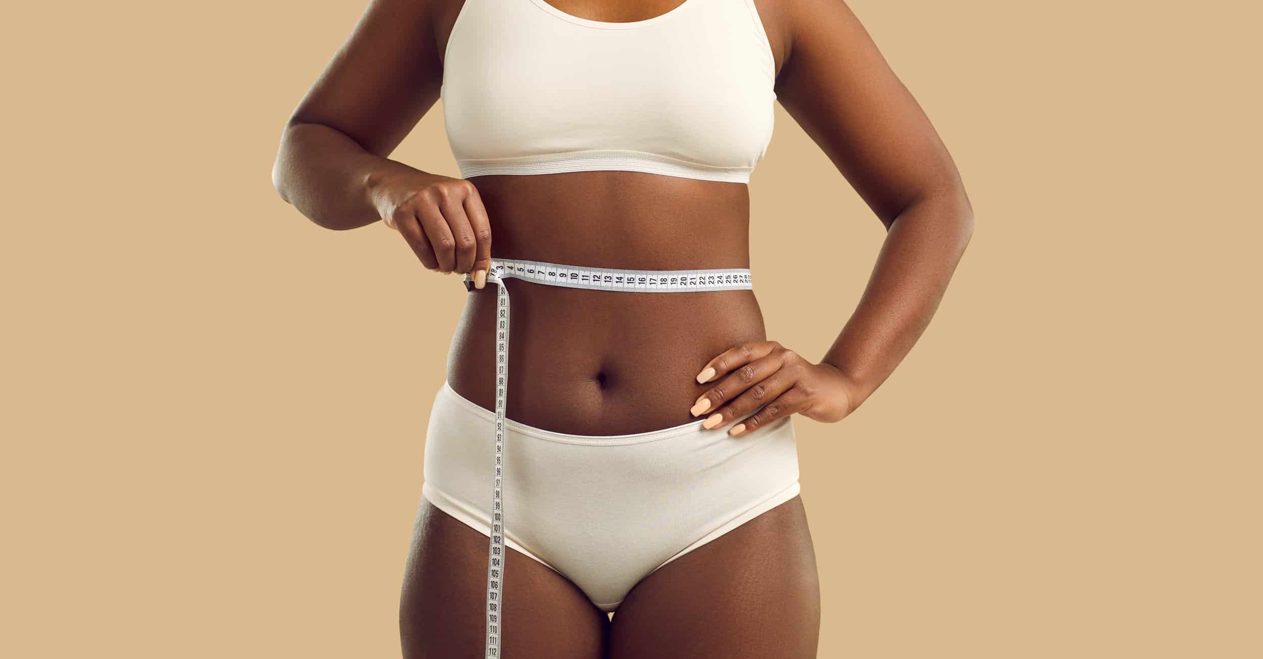 Plus size young black woman with belly fat measuring her waist with tape measure